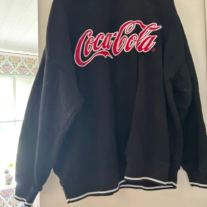 Svart Coca-Cola sweatshirt Divided S - Svart sweatshirt från H&M Divided med stor Coca-Cola-logga i rött och vitt på bröstet. Tröjan har långa ärmar, ribbade muddar och vita ränder vid ärmslut och nederkant. Perfekt för dig som gillar streetstyle och vill sticka ut.
