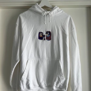 Vit hoodie med broderad Bluey-figur - Säljer en vit hoodie från Just Hoods i storlek M med broderad Bluey-figur på bröstet. Bluey och Bingo leker Grannies - Janet och Rita. Hoodien har snörning vid huvan, stor magficka och långa ärmar. Perfekt för dig som gillar chill och tecknade motiv.