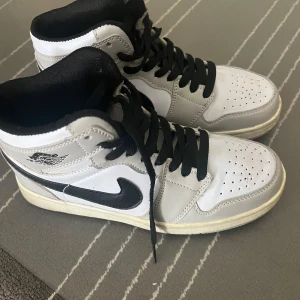 Nike Air Jordan 1 High vit/svart 37.5 - Snygga Nike Air Jordan 1 High i vitt och grått med svarta detaljer och klassisk svart swoosh. Skorna har svarta snören, perforerad tå och hög siluett. Tillverkade i skinn och syntet, perfekta för dig som gillar streetstyle och ikoniska sneakers. Lite skada finns men de inget som tål vatten när de regnar.
