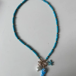 Turkost pärlhalsband med berlocker - Superfint halsband med små turkosa pärlor och gulddetaljer. Hängsmycken i form av en blå fisk, en stjärna och en vit pärla ger en lekfull vibe. Perfekt för dig som vill sticka ut med färg och coola detaljer. Hämta i Majorna eller ibland kan träffa i stan 