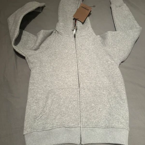Grå hoodie från Burberry med dragkedja - Snygg grå hoodie från Burberry med klassiskt rutigt foder i huvan. Tröjan har hel dragkedja framtill, fickor och ribbade muddar. Perfekt för dig som vill ha en stilren och exklusiv look med en touch av Burberrys ikoniska mönster.