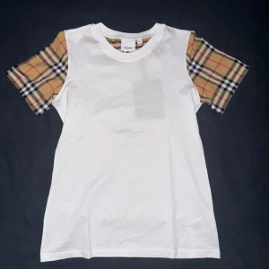 Vit Burberry t-shirt med rutiga ärmar - Snygg vit t-shirt från Burberry med klassiska rutiga ärmar i beige, svart, rött och vitt. T-shirten har rund halsringning och är tillverkad i mjuk bomull. Perfekt för dig som vill ha en stilren look med en touch av lyx.