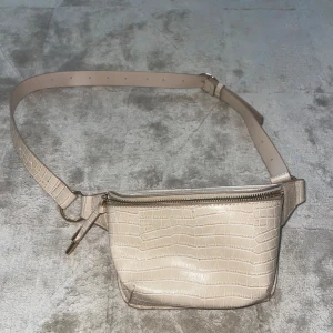 Beige magväska i crocopräglat skinn - Snygg beige magväska med crocopräglat mönster och justerbart bälte. Väskan har dragkedja i guld och stilren design som passar perfekt till streetwear eller när du vill ha händerna fria.