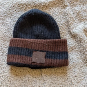 Randig mössa från Acne Studios - Snygg ribbstickad mössa från Acne Studios i svart och brunt med bred uppvikt kant och randigt mönster. Mössan har Acne Studios klassiska ansiktslogga framtill och är tillverkad i 100% ull. Perfekt accessoar för kyliga dagar.