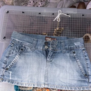 Ljusblå jeanskjol från Pershka - Säljer en snygg ljusblå jeanskjol från Pershka Denim i klassisk kort modell. Kjolen har slitna detaljer, fem fickor och bälteshällor. Baksidan har en vit patch med blommor och fjärilsmotiv. Perfekt för dig som gillar en chill och trendig stil.