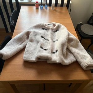 Vit teddyjacka från Gina Tricot M - Mysig vit teddyjacka från Gina Tricot i storlek M. Jackan har fluffigt teddy-material, snygga svarta spännen framtill och lång ärm. Perfekt för dig som gillar en chill och trendig look under höst och vinter. Defekt - spilt något blått på, vet tyvärr inte vad då det var så när ja köpte den på Plick därav priset 
