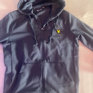 Svart hoodie från Lyle & Scott M - Svart hoodie från Lyle & Scott med dragkedja och huva. Klassisk gul logga på bröstet och snörning i huvan. Mjukt bomullsmaterial och ribbade muddar vid ärmslut och nederkant. Perfekt för chill dagar eller när du vill ha en clean style.