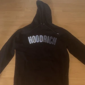 Svart hoodie från Hoodrich med tryck - Svart hoodie från Hoodrich i storlek M med stort, vitt och grått HOODRICH-tryck över bröstet. Tröjan har huva med snörning och är långärmad. Materialet är mjuk bomull, perfekt för chill dagar. Snygg streetwear-vibe och klassisk passform.