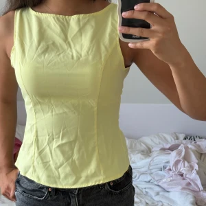 Gul ärmlös topp med knyt - Superfin ljusgul ärmlös linnetopp med figurnära passform och snygg knytning i ryggen. Toppen har en enkel, stilren design med rundad halsringning och slät yta. Perfekt att styla med jeans eller kjol för en fräsch look.