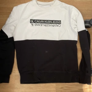 Svartvit sweatshirt från Calvin Klein Jeans - Snygg svartvit sweatshirt från Calvin Klein Jeans i storlek S. Tröjan har ett stilrent blockmönster med vit överdel och svarta ärmar och nederdel. Loggan är tryckt framtill i svart och vitt. Mjuk bomullskvalitet och rund hals. Perfekt för chill och streetwear.