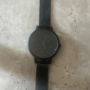 Svart klocka från Even&Odd med meshband - Stilren svart klocka från Even&Odd med rund urtavla dekorerad med små svarta stenar för en cool textur. Klockan har ett svart mesharmband i metall och minimalistisk design utan siffror. Perfekt accessoar för att lyfta din outfit. 