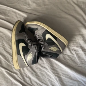 Nike Air Jordan 1 High grå/svarta - Snygga Nike Air Jordan 1 High i grått och svart med vita detaljer och klassisk vinge-logga på sidan. Skorna har snörning, hög siluett och den ikoniska swoosh-loggan. Perfekta för dig som vill sticka ut med streetstyle.