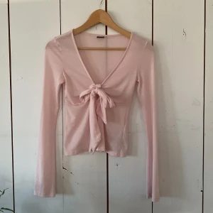 Ljusrosa omlottopp från Gina Tricot - Supersöt ljusrosa omlottopp från Gina Tricot i mjuk bomullsmix. Toppen har lång ärm, v-ringning och en snygg knytning framtill som ger en feminin touch. Perfekt att styla med jeans eller kjol för en trendig look.