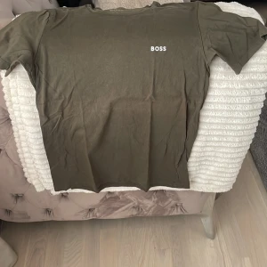 Olivgrön t-shirt från BOSS - Olivgrön t-shirt från BOSS med enkel vit logotyp på bröstet. Klassisk rund hals och korta ärmar. T-shirten är tillverkad i mjuk bomull och har en clean, stilren look som passar perfekt till jeans eller shorts.