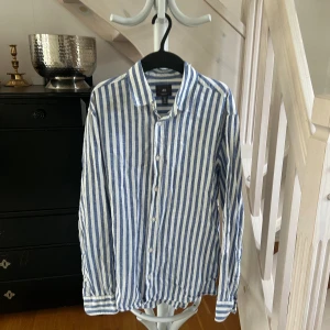 Randig blåvit skjorta från H&M - Snygg blåvit randig skjorta från H&M i regular fit. Skjortan har långa ärmar, klassisk krage och knäppning framtill. Tillverkad i ett lätt och luftigt linnematerial, perfekt för varmare dagar. Enkel och stilren design som passar till många outfits.