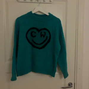 Turkos stickad tröja med hjärta - Turkos stickad tröja med svart hjärta och smiley på bröstet, samt bokstäverna CW. Tröjan har rund halsringning och långa ärmar. Perfekt för dig som gillar färg och vill sticka ut lite extra. 