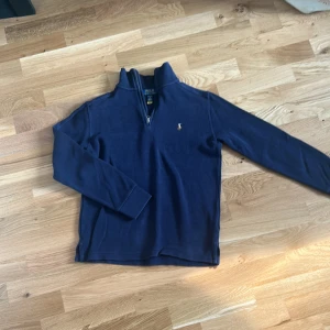 Marinblå half zip tröja Polo Ralph Lauren - Snygg marinblå långärmad tröja från Polo Ralph Lauren med half zip och klassisk broderad logga på bröstet. Tröjan är stickad i bomull och har en hög krage som ger en chill och stilren vibe. Perfekt för dig som gillar preppy stil och vill ha något som funkar året runt.