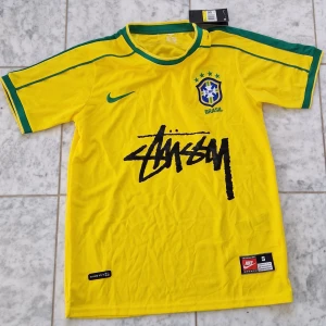 Brasilien x Stüssy x Nike gul fotbollströja - Gul Brasilien fotbollströja i samarbete med Stüssy och Nike. Tröjan har gröna detaljer på krage och ärmar, Brasilien-emblem på bröstet och Stüssy-logga i svart framtill. Tillverkad i lätt polyester, perfekt för match eller streetwear. Brazil x Stussy brazil t shirtz riktigt fet t-shirt som passar för alla. Storlek S. 