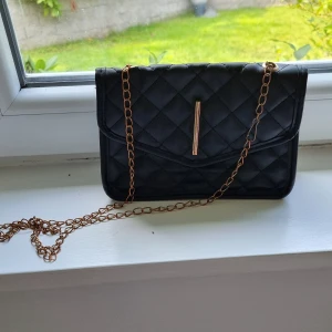 Svart quiltad axelväska med kedja - Snygg svart axelväska i quiltad design med guldfärgad kedjerem och stilren metall-detalj framtill. Perfekt storlek för mobilen och det viktigaste. Väskan har en klassisk look och passar till många outfits.