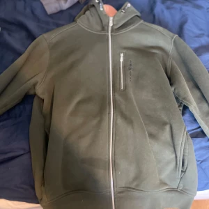 Grön hoodiejacka med dragkedja - Mörkgrön hoodiejacka med hel dragkedja framtill och en extra bröstficka med dragkedja. Jackan har huva med snörning och är långärmad. Perfekt för chill dagar eller när du vill ha en enkel streetstyle.