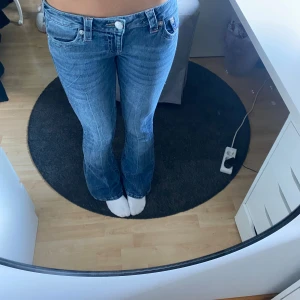 True Religion bootcut jeans blå, stl 26 - Säljer ett par snygga blå jeans från True Religion i klassisk bootcut-modell. Jeansen har låg midja, fem fickor och ikoniska bakfickor med kontrastsömmar. Tillverkade i mjukt denim med lätt slitning för en cool look.