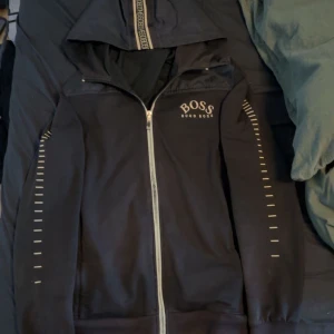 Svart hoodie från Hugo Boss - Svart hoodie från Hugo Boss med huva och dragkedja hela vägen. Snygga vita detaljer längs ärmarna och logga på bröstet. Hoodien har sportig vibe och är perfekt för dig som gillar stilrena plagg med en twist.