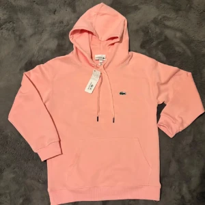 Ljusrosa hoodie från Lacoste - Snygg ljusrosa hoodie från Lacoste med klassisk krokodillogga på bröstet. Tillverkad för en skön och mjuk känsla. Perfekt för dig som gillar stilrena och bekväma plagg. Passar dig som är mellan 170-175.
