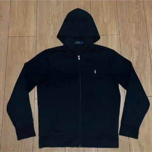 Svart hoodie från Polo Ralph Lauren - Svart hoodie från Polo Ralph Lauren med dragkedja och klassisk huva. Liten broderad logga på bröstet. Perfekt för en chill och stilren look. Passar dig som är mellan 185-190 perfekt.