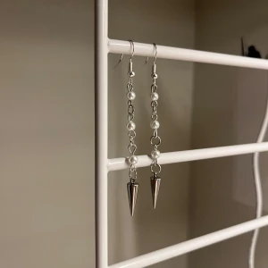 Silvriga örhängen med pärlor och spikes - Långa örhängen i silverfärgad metall med kedja dekorerad med vita pärlor och avslutande spetsiga spikes. Perfekt för dig som gillar edgy detaljer och vill sticka ut med din stil. Rostfritt stål och handgjort 