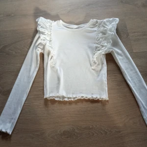 Vit ribbad topp med volang från Kappahl - Supersöt vit långärmad topp från Kappahl med ribbad struktur och volangdetaljer på axlarna. Rund hals och lätt croppad med vågig kant nedtill. Perfekt för dig som vill ha något lite extra💕Den är en liten rosa fläck på ryggen💕( finns katt i hemmet)