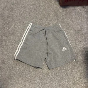 Grå shorts från Adidas - Snygga grå shorts från Adidas med klassiska vita ränder längs sidorna. De har en bekväm resårmidja med snörning för justerbar passform. Perfekta för en avslappnad stil eller sportig look.