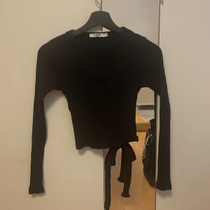 Svart ribbad topp från NA-KD - Snygg svart ribbad topp från NA-KD med långa ärmar och öppen rygg med knytning i ryggen. Är använd en gång. Säljer då den är för liten