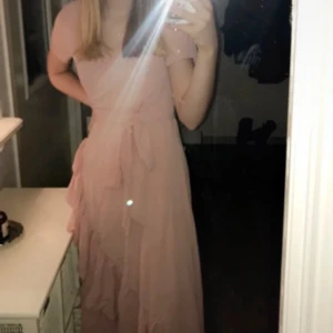 Rosa långklänning med volangdetaljer - Elegant rosa långklänning med korta ärmar och volangdetaljer. Klänningen har en knytning i midjan. Använd på en babyshower och på ett dop .