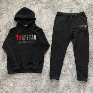 Svart hoodie och byxor från Trapstar - Säljer ett svart Trapstar-set med hoodie och matchande byxor. Hoodien har en stor logotyp fram och justerbar huva med dragsko. Byxorna har samma logotyp på benet och en elastisk midja med dragsko. Perfekt för en avslappnad stil!