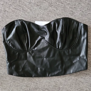 Svart skinnliknande korsett - Snygg svart korsett i skinnliknande material med dragkedja bak. Perfekt för en edgy look. Den är axelbandslös och har en stilren design som passar till många olika outfits.