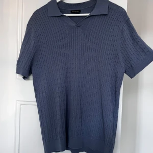 Blå pikétröja från Massimo Dutti - Snygg blå pikétröja från Massimo Dutti i bomull och linne. Tröjan har korta ärmar och ett diskret ribbat mönster. Perfekt för en stilren look. Hör av dig vid minsta fråga eller fundering!