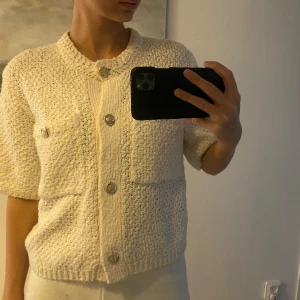 Beige stickad kofta med knappar - Säljer en snygg beige stickad kofta med korta ärmar och dekorativa knappar framtill. Perfekt för en stilren look. Passar bra till både jeans och kjol.