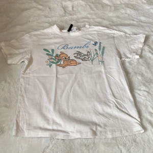 Vit Bambi t-shirt från H&M Divided - Säljer en söt vit t-shirt från H&M Divided med ett Bambi-tryck i pastellfärger. T-shirten har korta ärmar och är gjord i mjuk bomull. Perfekt för alla Disney-fans! 🦌