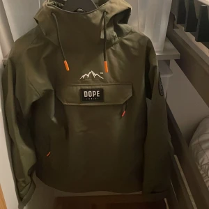 Grön jacka från Dope - Säljer en grön jacka från Dope med en stor ficka framtill och justerbar huva. Jackan har en broderad logga på bröstet och en patch på ärmen. Perfekt för utomhusaktiviteter med stil! 💚