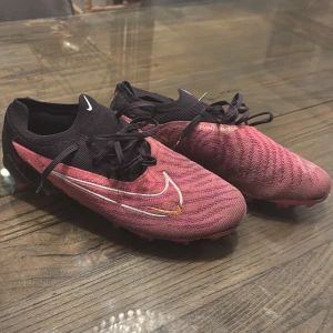 Nike Phantom fotbollsskor - Snygga Nike Phantom fotbollsskor i rosa och svart med snörning. Skorna har en modern design med tydliga Nike-loggor och är perfekta för fotbollsplanen.