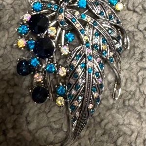 Glittrig brosch med färgglada stenar - En fantastisk glittrig brosch i form av ett löv, dekorerad med gnistrande stenar i blått, rosa och lila. Perfekt för att ge en extra touch av glamour till din outfit.