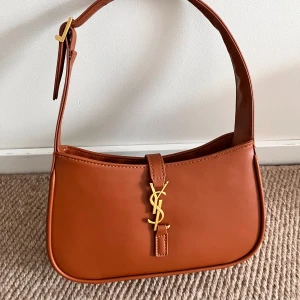 Brun axelväska YSL - Snygg brun axelväska i guldiga detaljer. Väskan har en justerbar axelrem och ett elegant YSL-emblem på framsidan. Jag har fått den i present och vet därför inte om den är äkta. Skicka dm för frågor/flera bilder