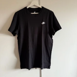 Svart t-shirt från Nike - Säljer en klassisk svart t-shirt från Nike med deras ikoniska logga i vitt på bröstet. Perfekt för en stilren och sportig look. T-shirten är kortärmad och tillverkad i mjukt bomullsmaterial. Knappt använd. Nypris 299kr