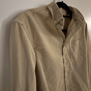 Skjorta - Säljer en stilren beige skjorta. Skjortan har långa ärmar och en klassisk krage med knappar. Perfekt för en avslappnad men ändå uppklädd look. 