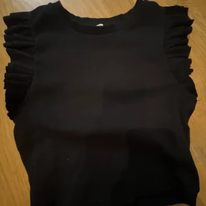 Svart ärmlös topp från Zara - Säljer en stilren svart ärmlös topp från Zara med volangdetaljer vid axlarna. Perfekt för att ge en extra touch till din outfit. Toppen är i ett mjukt material som känns skönt mot huden.