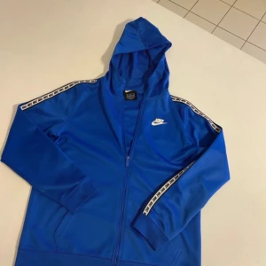 Blå huvtröja från Nike - Säljer en blå huvtröja från Nike med dragkedja och huva. Tröjan har vita detaljer längs ärmarna och en liten Nike-logga på bröstet. Perfekt för en sportig look!