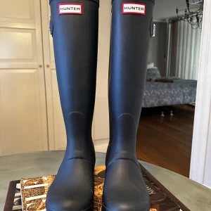 Svarta stövlar från Hunter - Snygga blå stövlar från Hunter med klassisk design och justerbart spänne på sidan. Perfekta för regniga dagar och håller fötterna torra och bekväma. Insidan har ett mönstrat foder för extra stil.