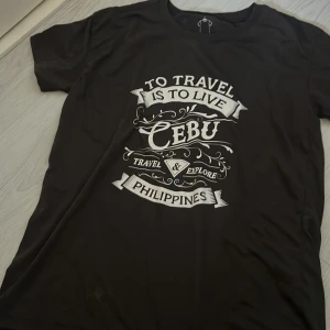Svart t-shirt från Kamisa - Säljer en svart t-shirt från Kamisa med tryck 'To Travel is to Live, Cebu, Philippines'. Tillverkad i polyester och bomull, perfekt för resor och äventyr.