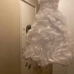 Vit klänning med volanger - Elegant vit klänning med volanger och detaljerad överdel. Klänningen har en vacker struktur med rynkade detaljer och en bred midjeband. Perfekt för speciella tillfällen.