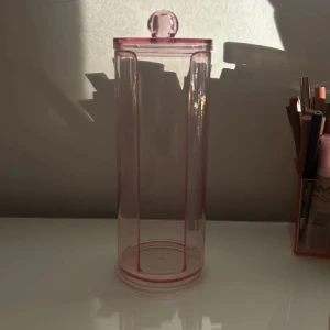 Rosa transparent behållare med lock - Säljer en stilren och praktisk rosa transparent behållare med ett runt lock. Perfekt för förvaring av småsaker eller sminkborstar. Den har en enkel och elegant design som passar in i de flesta rum.